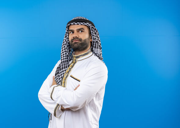 传统身着传统服装的阿拉伯男子站在蓝色的墙上 自信地看着一边 双手交叉放在胸前阿拉伯语男人旁边