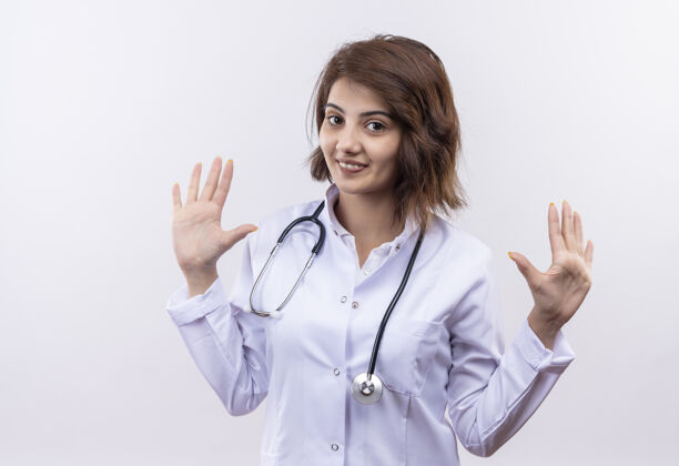 医生身穿白大褂 手持听诊器 微笑着举手投降的年轻女医生站在白墙上微笑站起来听诊器