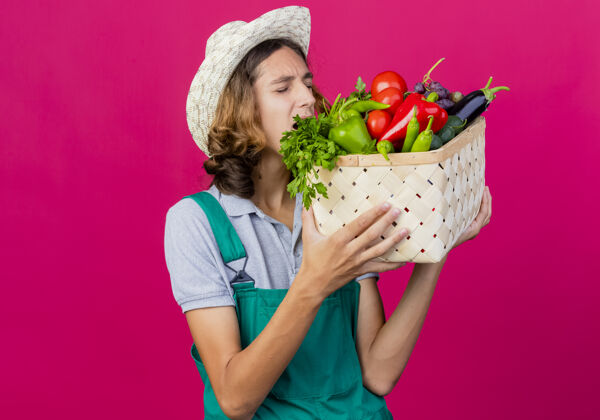 持有年轻的园丁穿着连体衣戴着帽子拿着装满新鲜蔬菜的箱子测试蔬菜年轻