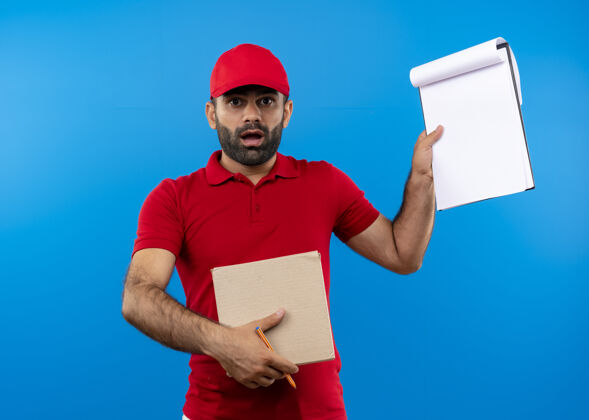 送货有胡子的送货员穿着红色制服 戴着帽子 拿着一个盒子包裹 上面写着一个看起来很惊讶的剪贴板 站在蓝色的墙上帽子惊喜制服