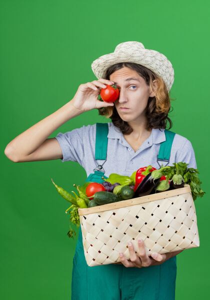 帽子年轻的园丁穿着连体衣戴着帽子拿着装满新鲜蔬菜的箱子穿番茄立场