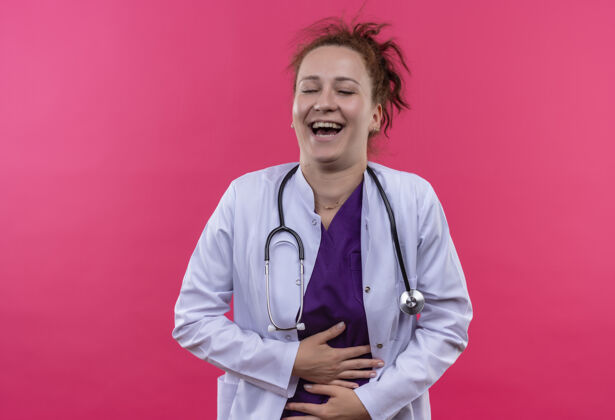 外套年轻的女医生穿着白大褂 手持听诊器 站在粉红色的墙上 笑着摸着她的肚子摸笑站
