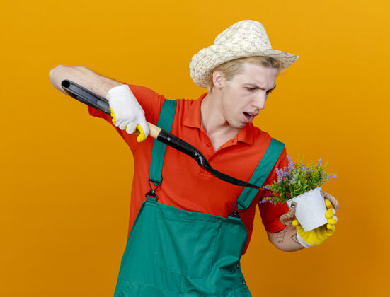 连身衣年轻的园丁穿着连体衣戴着帽子拿着铲子和盆栽植物罐子拿着帽子