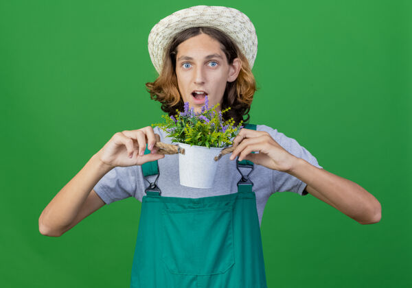 穿年轻的园丁穿着连体衣戴着帽子拿着盆栽植物惊讶帽子年轻人花园