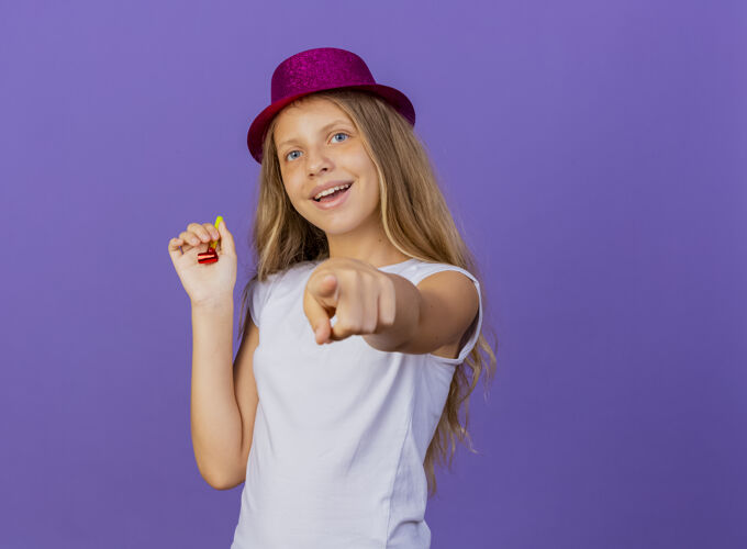 快乐戴着节日礼帽的漂亮小女孩用食指指着镜头微笑着快乐而积极 生日派对的概念站在紫色的背景上微笑小女孩