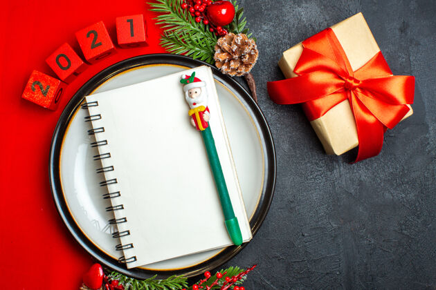 圣诞上图为新年背景 餐盘上有螺旋形笔记本装饰配件 红色餐巾上有杉木树枝和数字 黑色桌子上有红丝带礼物笔记本杉木树枝