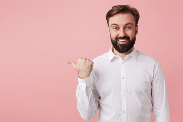 男性快乐迷人的年轻黑发男性 胡子浓密 身穿白衬衫 在粉红色的墙上摆姿势 展示他愉快的情绪 并用拇指指向一边郁郁葱葱朋友短裤