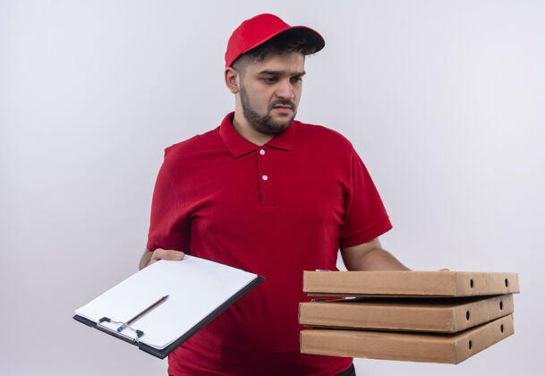 站年轻的送货员身穿红色制服 头戴鸭舌帽 手里拿着比萨饼盒和剪贴板 手里拿着钢笔和空白页 看上去困惑不解 试图做出选择剪贴板钢笔帽子