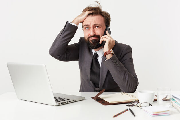 电话紧张的黑胡子男人坐在工作台上紧张地打电话 头发乱蓬蓬的 脸上一副迷惑不解的样子 穿着灰色西装男人漂亮拍摄