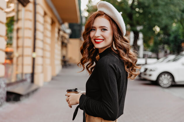 年轻戴着贝雷帽的姜黄色女人回头看长发微笑的女孩在街上喝咖啡秋天肖像咖啡