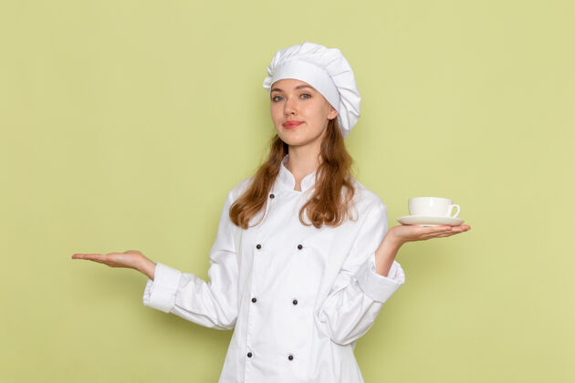 女厨师身着白色厨师服的女厨师正对着绿色墙壁微笑着拿着白色咖啡杯抱着前面淋浴帽