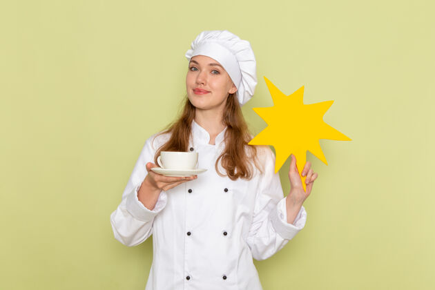 肖像女厨师身穿白色厨师服 手持黄色标牌 绿色桌子上放着一杯咖啡 厨房烹饪美食 女性颜色快乐女厨师咖啡