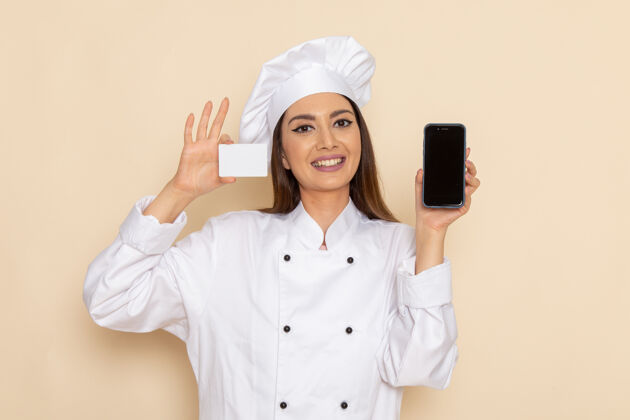 女性身穿白色厨师服的年轻女厨师的正视图 在浅白的墙上拿着名片和电话前面电话举行