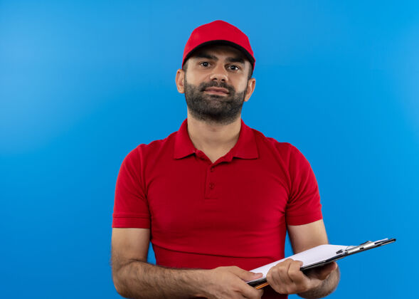 页面留着胡须的送货员穿着红色制服 戴着帽子拿着空白页的剪贴板 自信地微笑着站在蓝色的墙上制服站着微笑