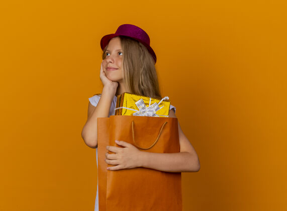 聚会戴着节日礼帽的漂亮小女孩拿着带礼物的纸袋 开心地看着一旁 橙色背景下站着生日派对的概念举行脸生日