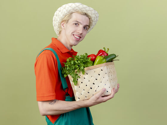 蔬菜年轻的园丁穿着连体衣 戴着帽子 手里拿着装满蔬菜的箱子 站在明亮的背景下 面带微笑地看着摄像机看穿男人
