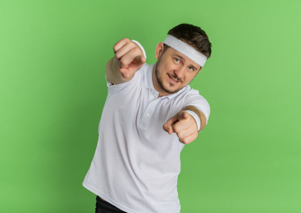 微笑身穿白衬衫 头戴头巾的年轻健身男子站在绿色的墙上 用食指指着你 脸上带着微笑教练年轻运动