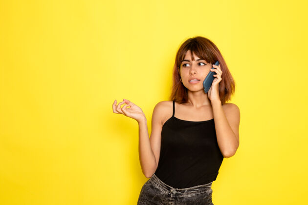 模特黄色墙壁上穿着黑色衬衫和灰色牛仔裤的年轻女性在讲电话的前视图牛仔裤年轻性感