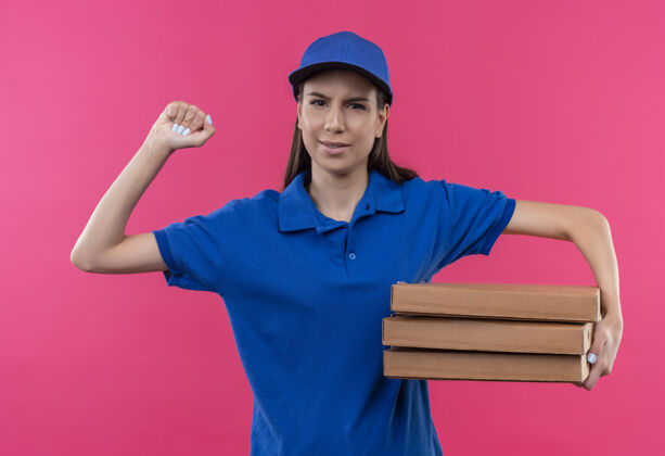 制服身穿蓝色制服 戴着帽子的年轻送货女孩举着一叠比萨饼盒 举起拳头严肃地看着镜头女孩堆着粉色