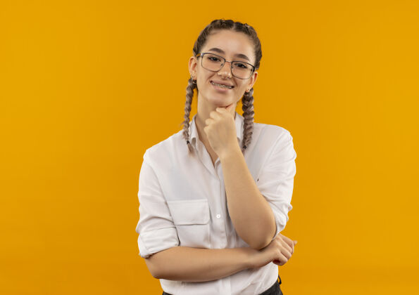 表情戴着眼镜 梳着辫子 穿着白衬衫的年轻女学生站在橙色的墙上 带着自信的微笑望着前方人学生站立
