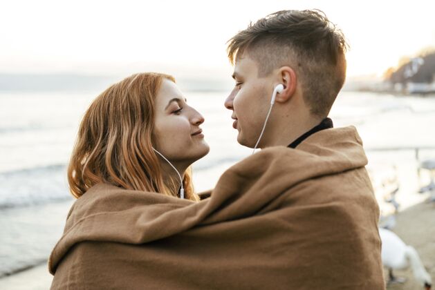 业余爱好冬天 一对年轻夫妇在沙滩上用耳机听音乐休闲探索户外