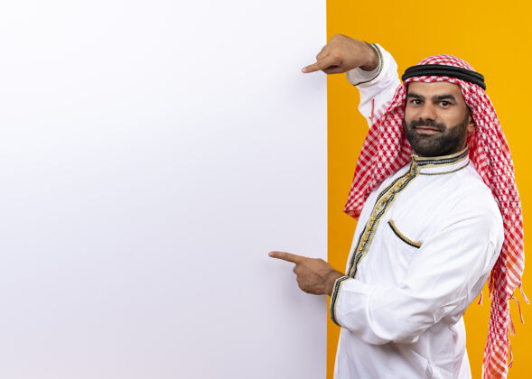 空白身着传统服装的阿拉伯商人指着空白的广告牌 自信地站在橙色的墙上看站穿