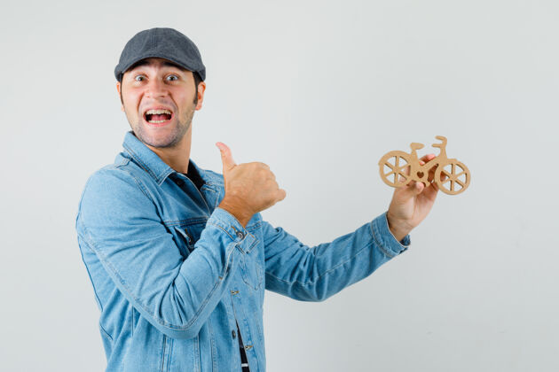 商人年轻人竖起大拇指 拿着一辆木制玩具自行车 穿着t恤 夹克 帽子 看上去很开心正面图肖像前面抱着