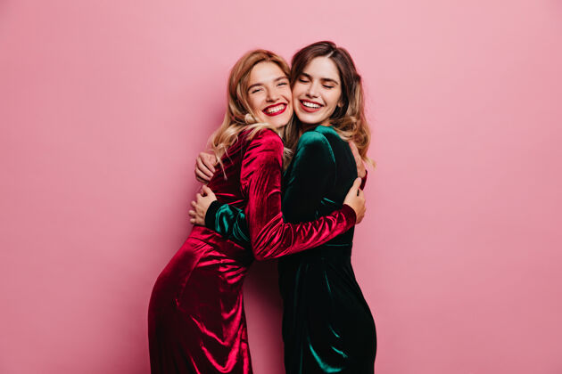 微笑兴奋的女性朋友在粉红色的墙上拥抱一些快乐的成年姐妹微笑着摆姿势的室内照片放松女人时尚