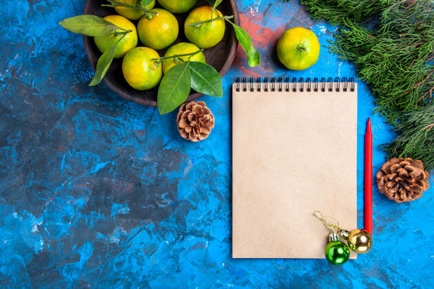 碗顶视图黄色的橘子和树叶在木制的碗一个笔记本和红色铅笔圣诞装饰品在蓝色的表面史密斯奶奶柠檬视图