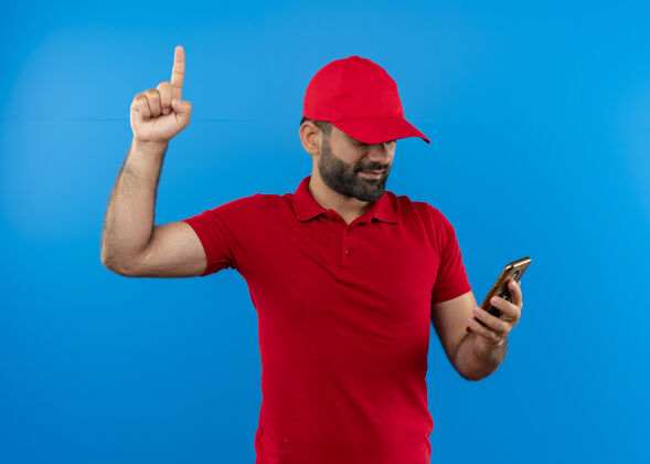 男人留着胡子的送货员穿着红色制服 戴着帽子 看着手机屏幕上的食指自信地站在蓝色的墙上移动信心看