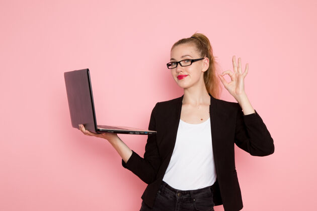 商务身穿黑色夹克的女上班族在浅粉色墙上手持和使用笔记本电脑的正面图严格正面微笑