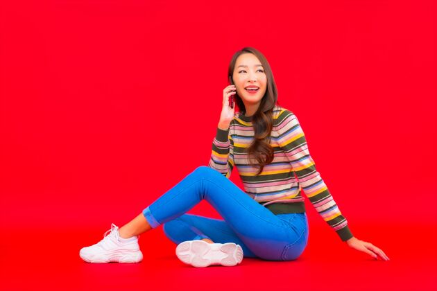 微笑肖像美丽的亚洲年轻女子在红色隔离墙上使用智能手机短信手机购买