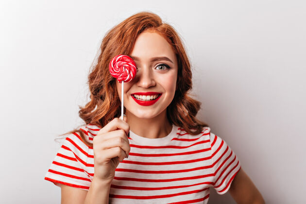 波浪发快活的姜女拿着红糖的特写照片快乐的欧洲卷发女孩在吃棒棒糖年轻微笑卷曲
