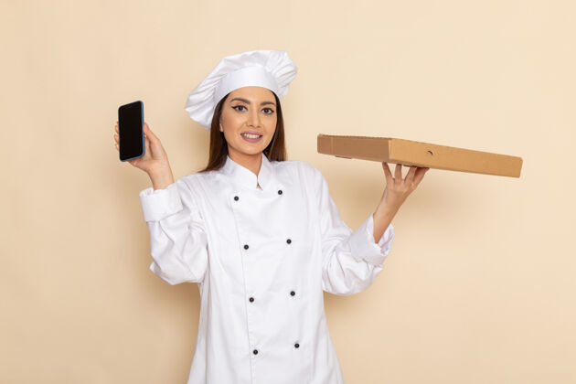 专业身穿白色厨师服的年轻女厨师正拿着智能手机和食物盒站在白色墙上厨师工人护士