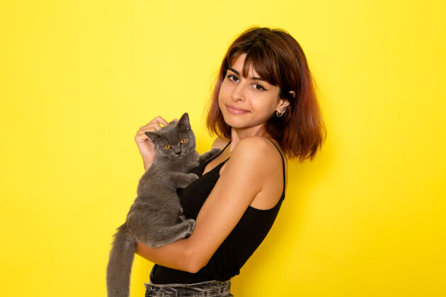 衬衫身穿黑色衬衫和灰色牛仔裤的年轻女性的正面视图 黄色墙上抱着灰色小猫漂亮女孩年轻