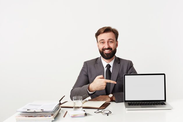 爱摄影棚拍摄的一个留着短胡子的黑发男人穿着正式的衣服在白色的墙上摆姿势 举着手在笔记本电脑上展示 兴高采烈地看着前面短裤坐着深色