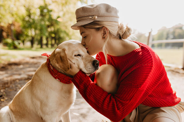 柔软美丽的金发女孩在秋天阳光明媚的公园亲吻她的可爱的狗穿着红色毛衣和时髦帽子的时髦年轻女子温柔地抱着宠物金发时尚年轻