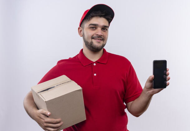 展示年轻的送货员穿着红色制服 戴着帽子拿着盒子包 展示着智能手机 自信地微笑着看着相机拿着帽子移动
