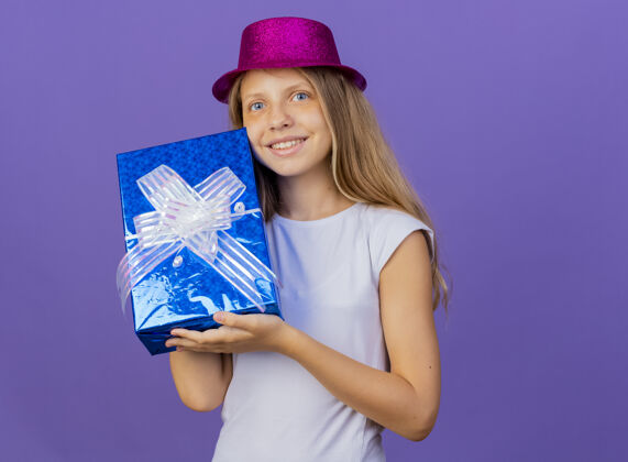 紫色美丽的小女孩戴着节日礼帽拿着礼盒看着相机笑脸盈盈 生日派对概念站在紫色的背景下微笑聚会快乐