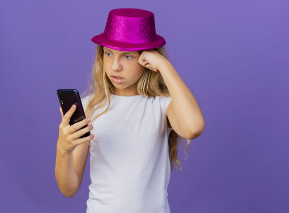 非常戴着节日帽子的漂亮小女孩用智能手机看着困惑和焦急 生日派对的概念站在紫色的背景上站小帽子