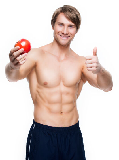 营养年轻快乐的健美运动员手拿苹果 竖起大拇指的画像-隔离在白墙上躯干健美微笑