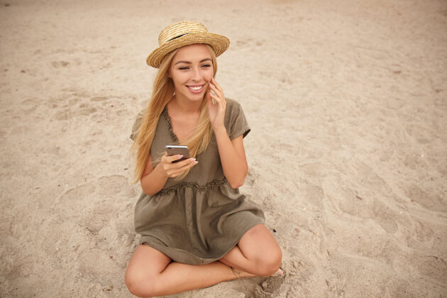 女士积极的金发长发美女盘腿坐在沙滩上 手里拿着智能手机 笑容满面 用举起的手抚摸着自己的脸手机长智能手机