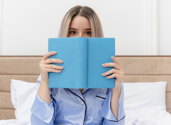 床穿着蓝色睡衣的年轻漂亮女人坐在床上 藏着书的脸在卧室内部的灯光背景下偷看女人漂亮室内