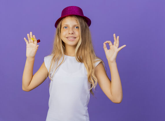 节日戴着节日礼帽的漂亮小女孩看着相机微笑着展示ok标志 紫色背景下站着生日派对的概念站生日派对