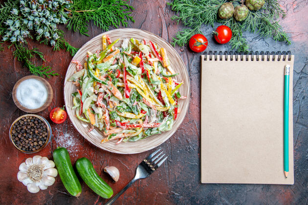松树顶视图蔬菜沙拉在盘子叉子上西红柿松枝黄瓜大蒜铅笔在记事本上在深红色的桌子上一餐食物大蒜