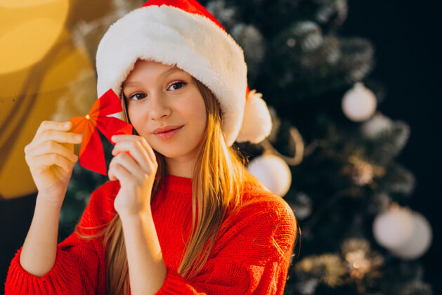 美丽可爱的少女戴着圣诞树旁的红色圣诞帽庆祝人帽子