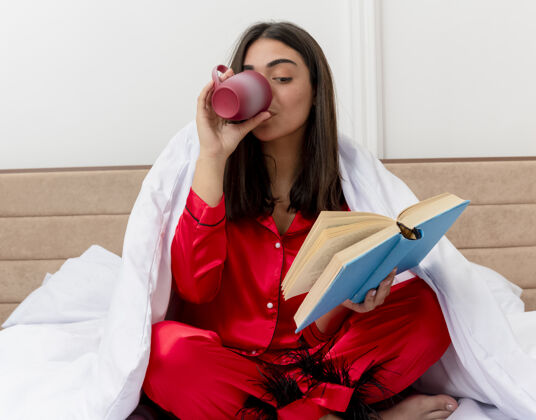 室内穿着红色睡衣的年轻漂亮女人坐在床上 裹着毛毯 拿着书 在卧室里喝着咖啡 背景很浅喝酒卧室女人