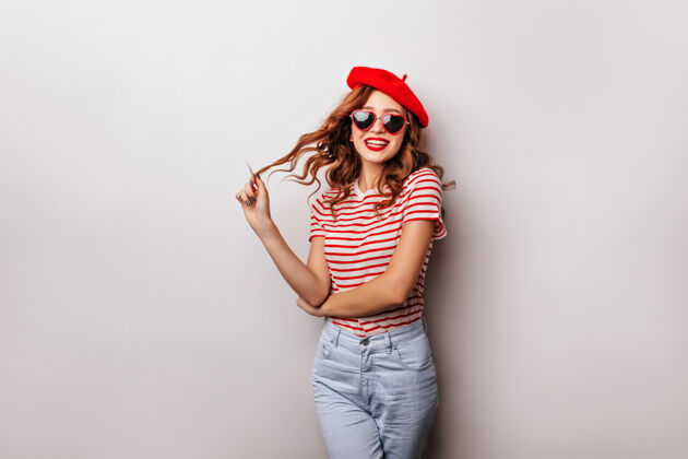 红发可爱的白人女孩穿着牛仔裤和贝雷帽站在白色的墙上室内照片笑着和蔼可亲的姜黄色头发的年轻女子肖像法国法国