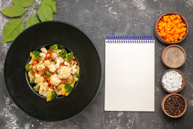 煎锅俯视图西兰花和花椰菜沙拉在黑碗不同的香料和削减胡萝卜碗笔记本在黑暗的表面香料顶部厨房用具