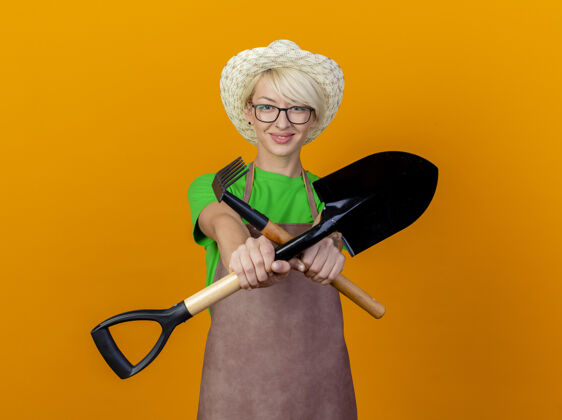 花园一位年轻的园丁 围裙上留着短发 戴着帽子 手里拿着小耙子和铲子 站在橙色的背景上微笑着看着相机女人迷你看